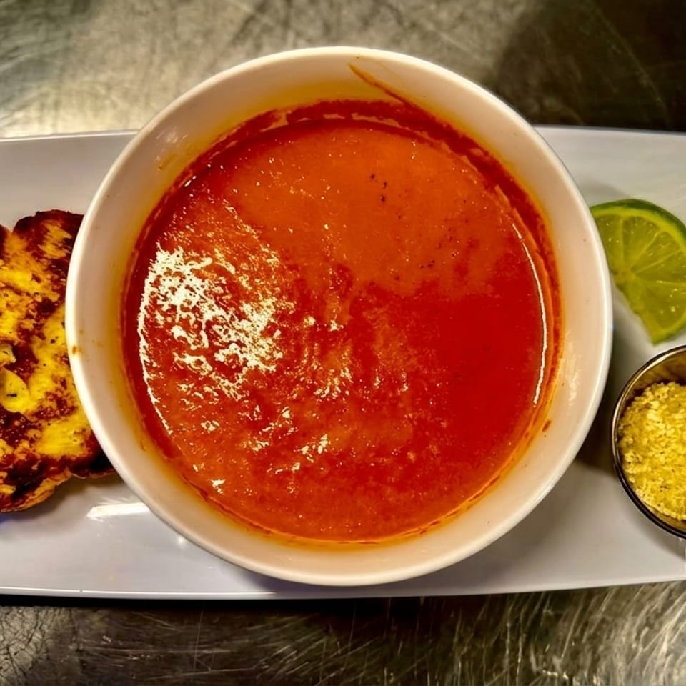 A bowl of tomato soup with a side of grilled cheese, lime, and parmesan cheese.